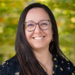 Woman with glasses and shoulder length brown hair in a black dress smiles at camera