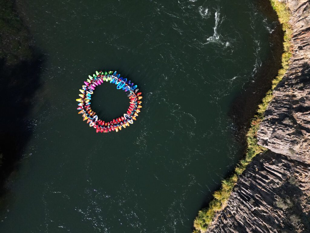 Aerial view of colorful kayaks in a circle on Klamath River next to a cliffside. 