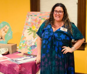 A woman in a blue dress poses at a table with artwork on display.