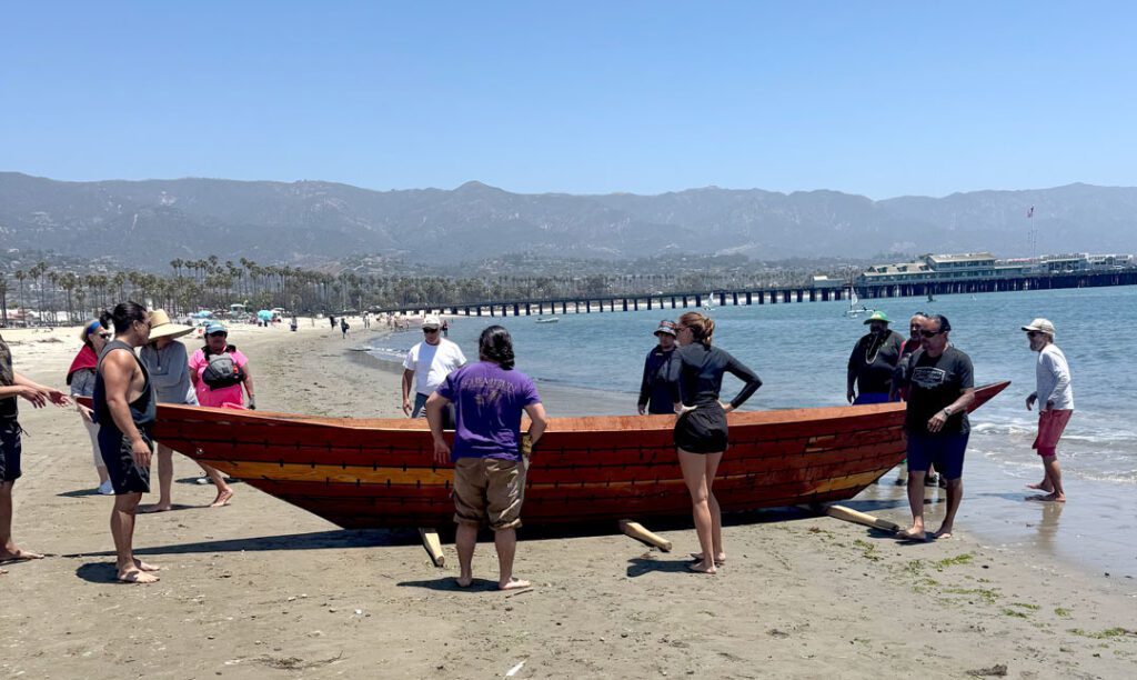 28-30a_NCF-Boat-web A group of people on a beach surround a simple wooden boat, known as a tomol.