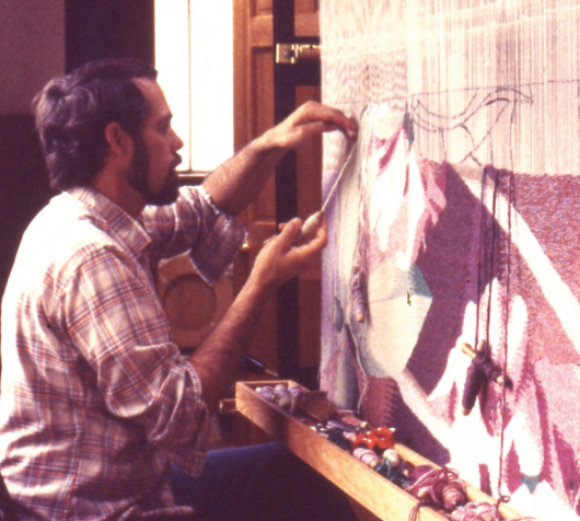 Middle aged white man with brown hair sits to the left of a painting he is working on.
