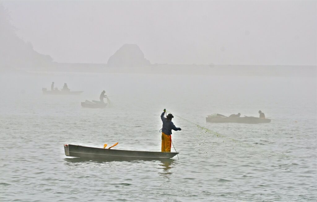 A Yurok fisherman stands on a boat pulling up a fishing net. Three boats with additional fishermen are in the background obscured by fog.