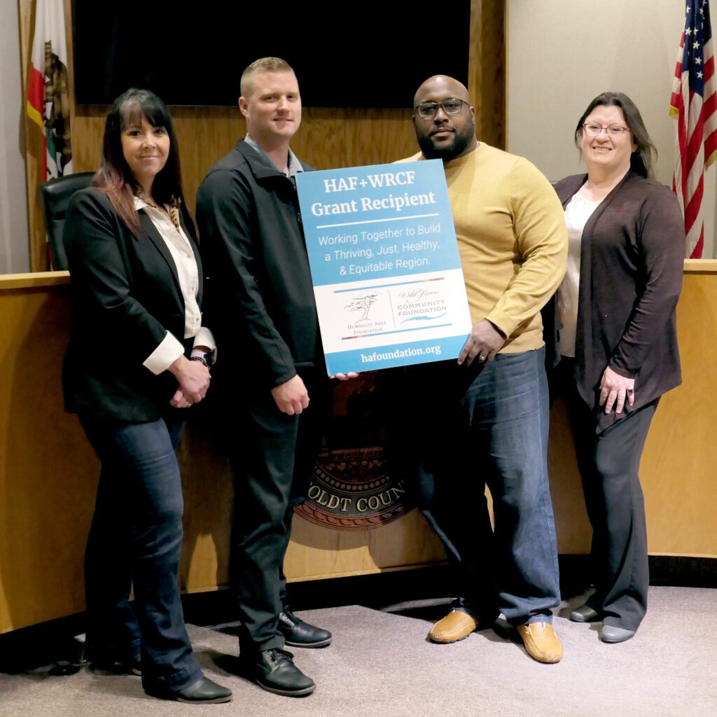 Four adults stand in a line in a board room and two of the adults hold a teal sign that reads HAF + WRCF Grant Recipient