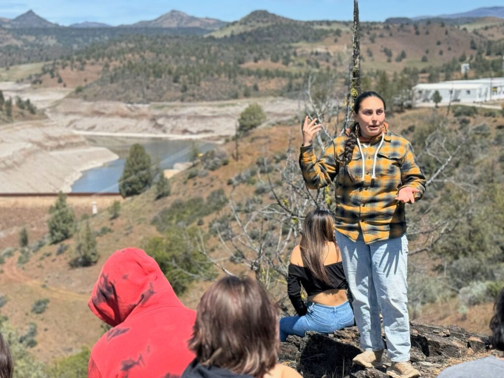 Person in yellow plaid and jeans speaks to crowd in front of Klamath River