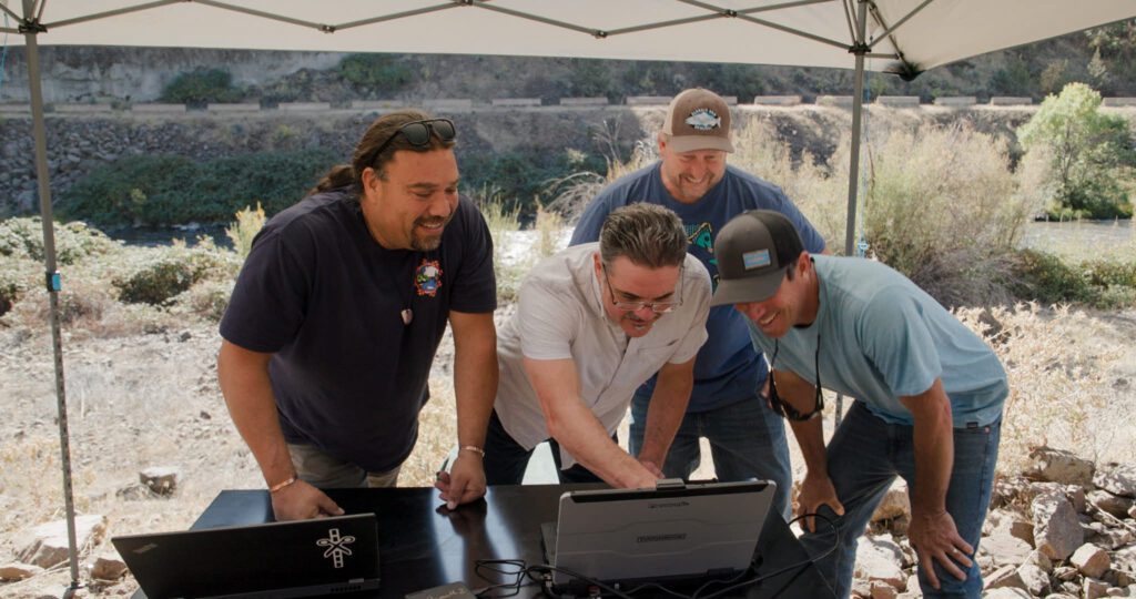 Four smiling people gather around a laptop outside