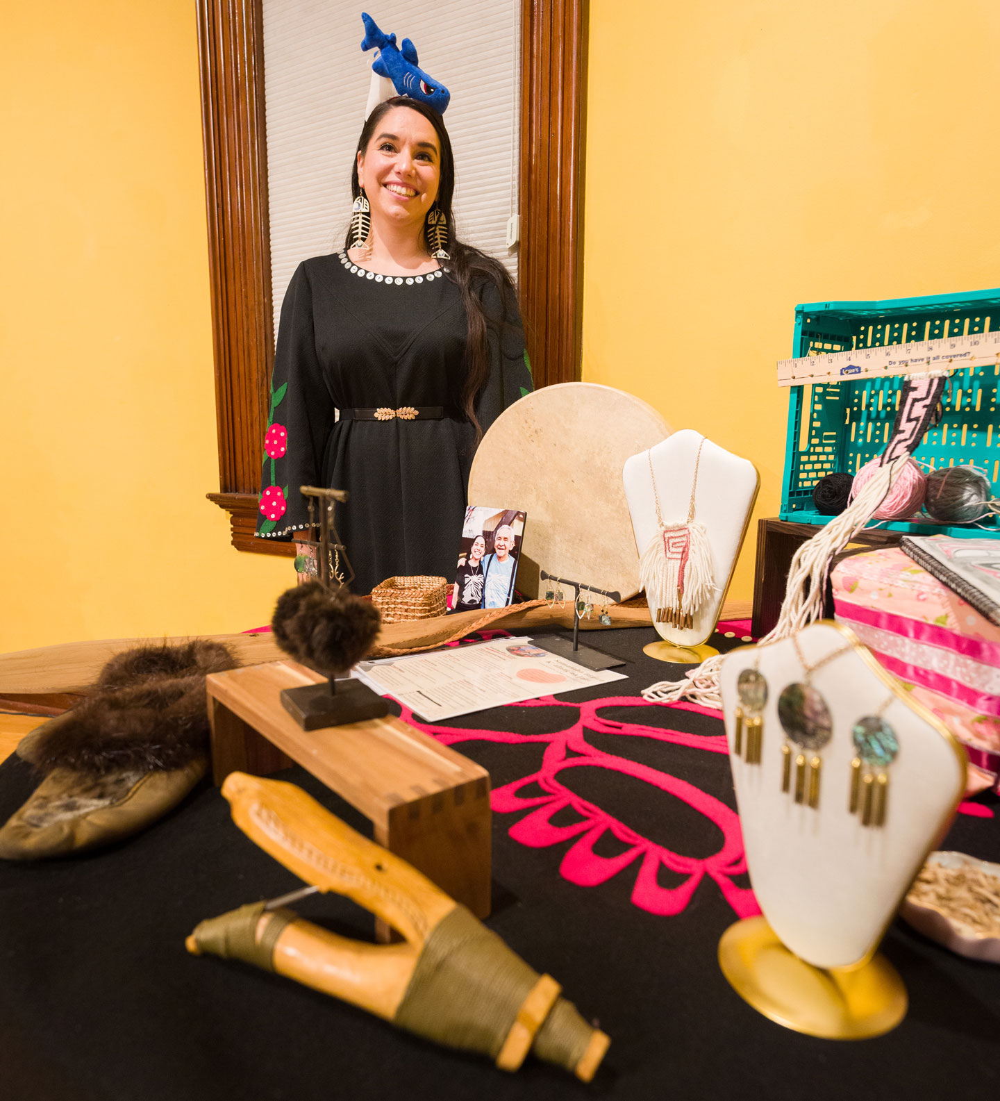 A woman poses at a table with various art pieces, including jewelry, textiles, and toys.