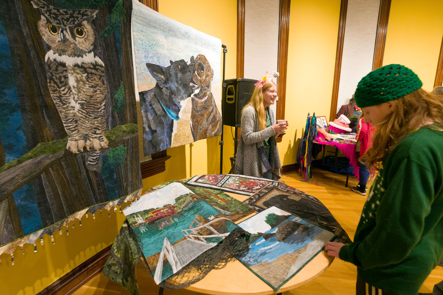 A woman stands in front of handmade quilts while a person in the foreground examines the quilts up close.